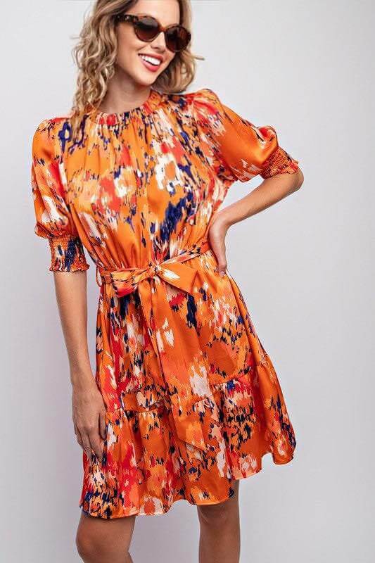 Woman wearing Autumn Radiance Dress by L Love, in amber with smocked sleeves, belted waist, ruched mock neck, and abstract print.