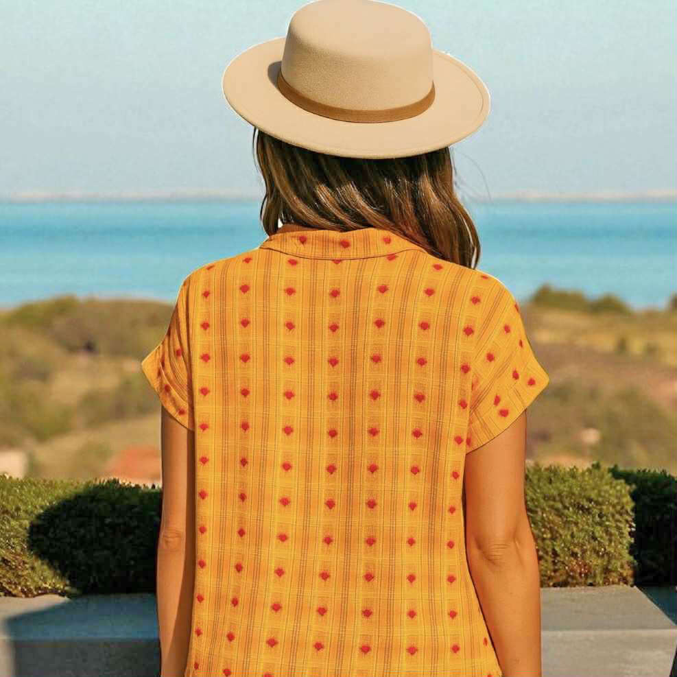 Back view of woman in yellow Hello Happiness Top by Umgee, featuring heart motifs and a sun hat against a scenic background.