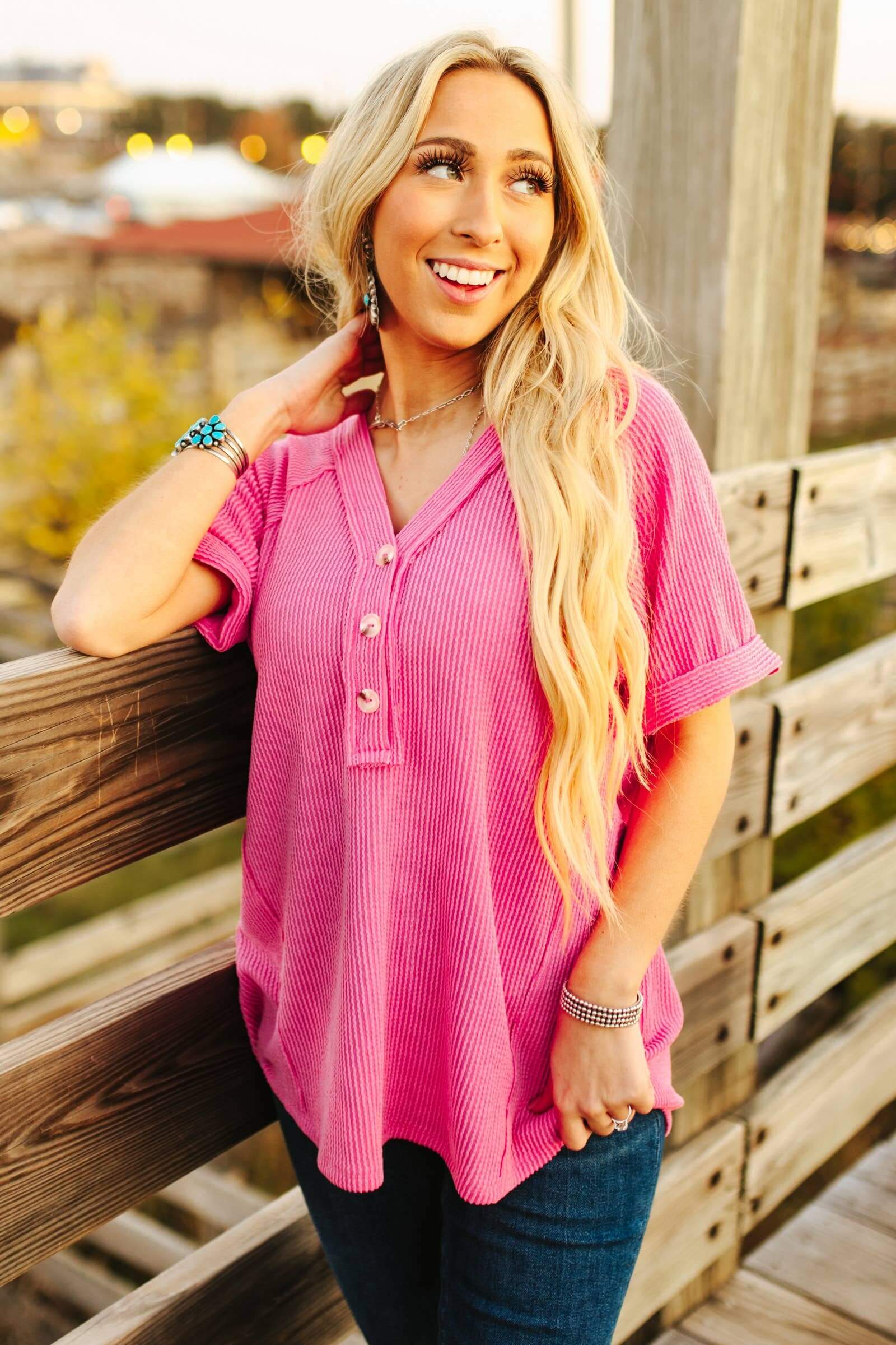 Woman wearing a pink shirred knit blouse with button details, showcasing a relaxed fit against a scenic wooden backdrop.