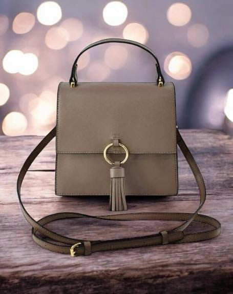 Taupe crossbody bag with ring design, tassel detail, and gold-tone hardware. Vegan leather bag with detachable strap on wood surface.