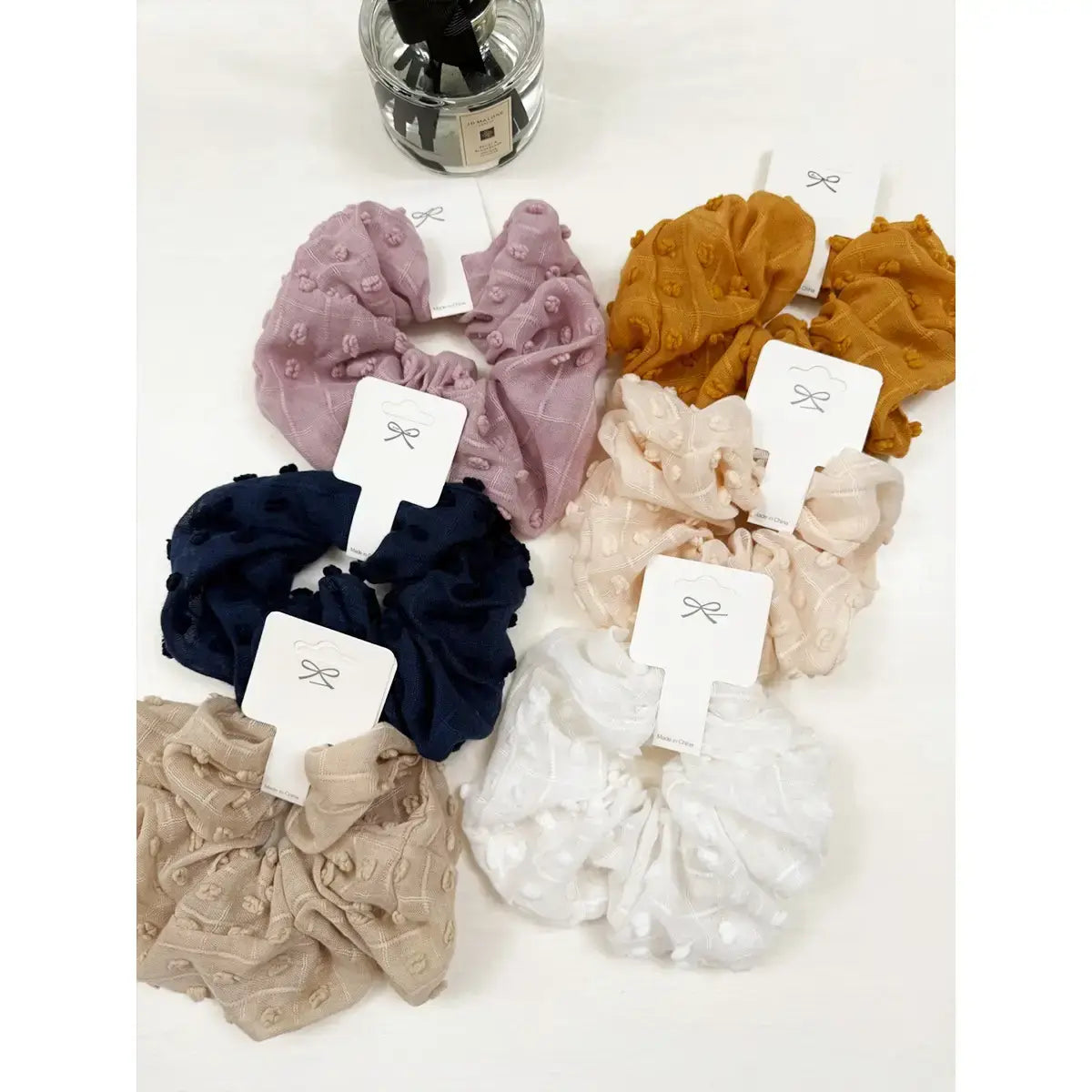 Assorted Cloud Comfort Scunchies in six colors: pink, mustard, navy, beige, cream, and white, showcasing plush pompom texture.