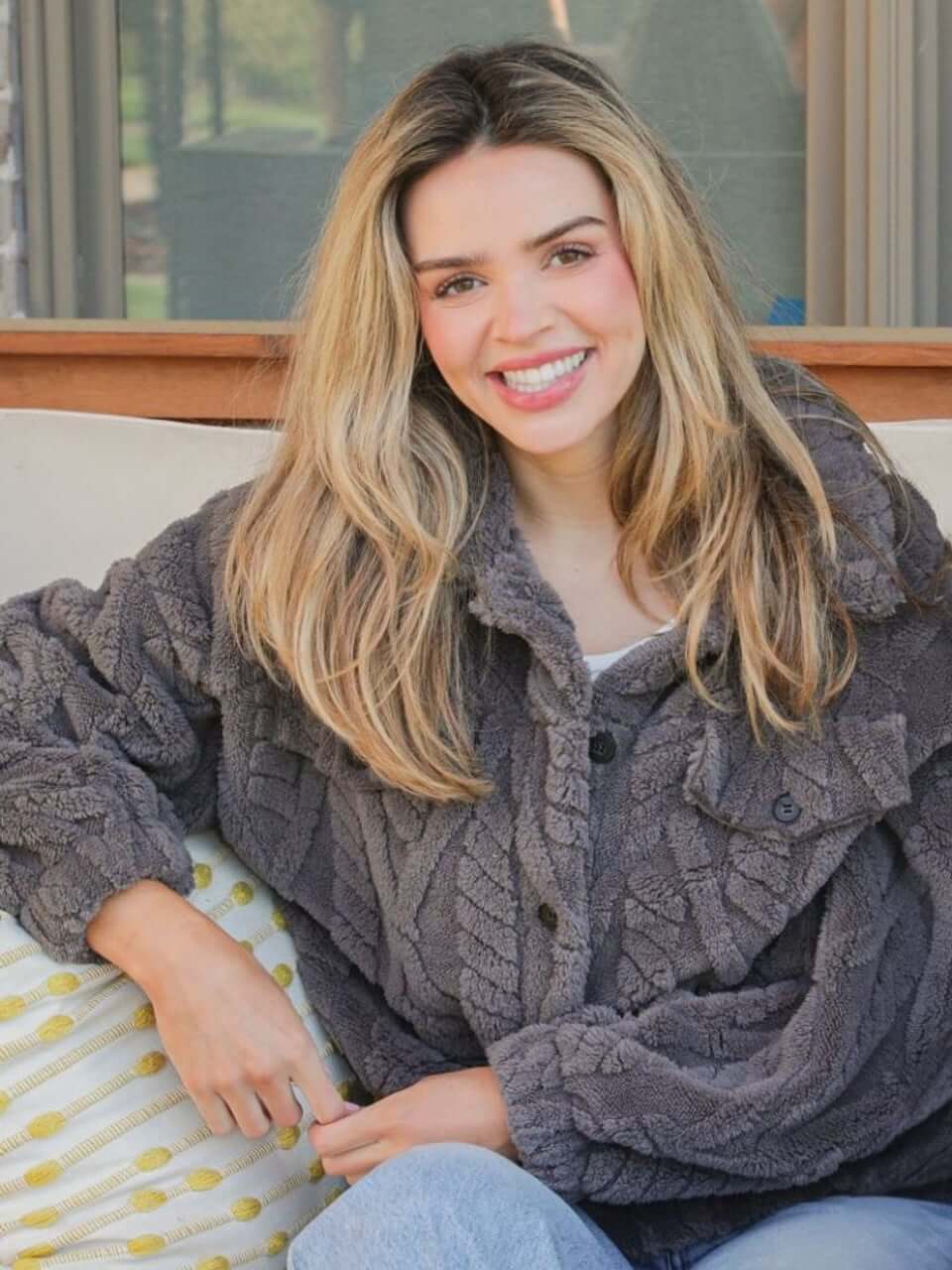 Woman wearing Simply Soft Sherpa Jacket in charcoal with a button front and cable knit design, seated comfortably and smiling.