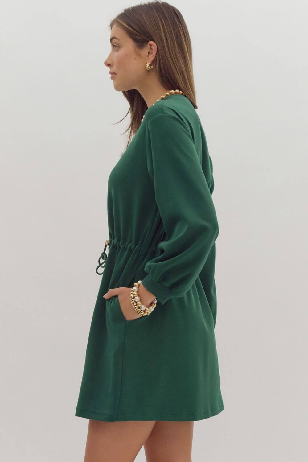 Woman wearing a dark green EVERGREEN BREEZE dress, featuring a crewneck and drawstring waist, side pockets, and long sleeves.