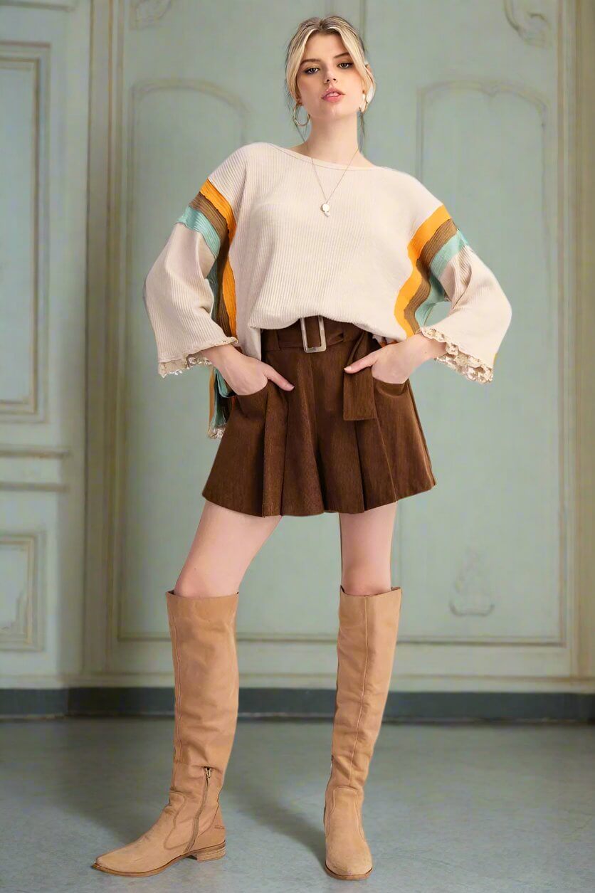 Woman in a casual outfit featuring a striped rib knit top, brown pleated skirt, and knee-high beige boots in a stylish pose.