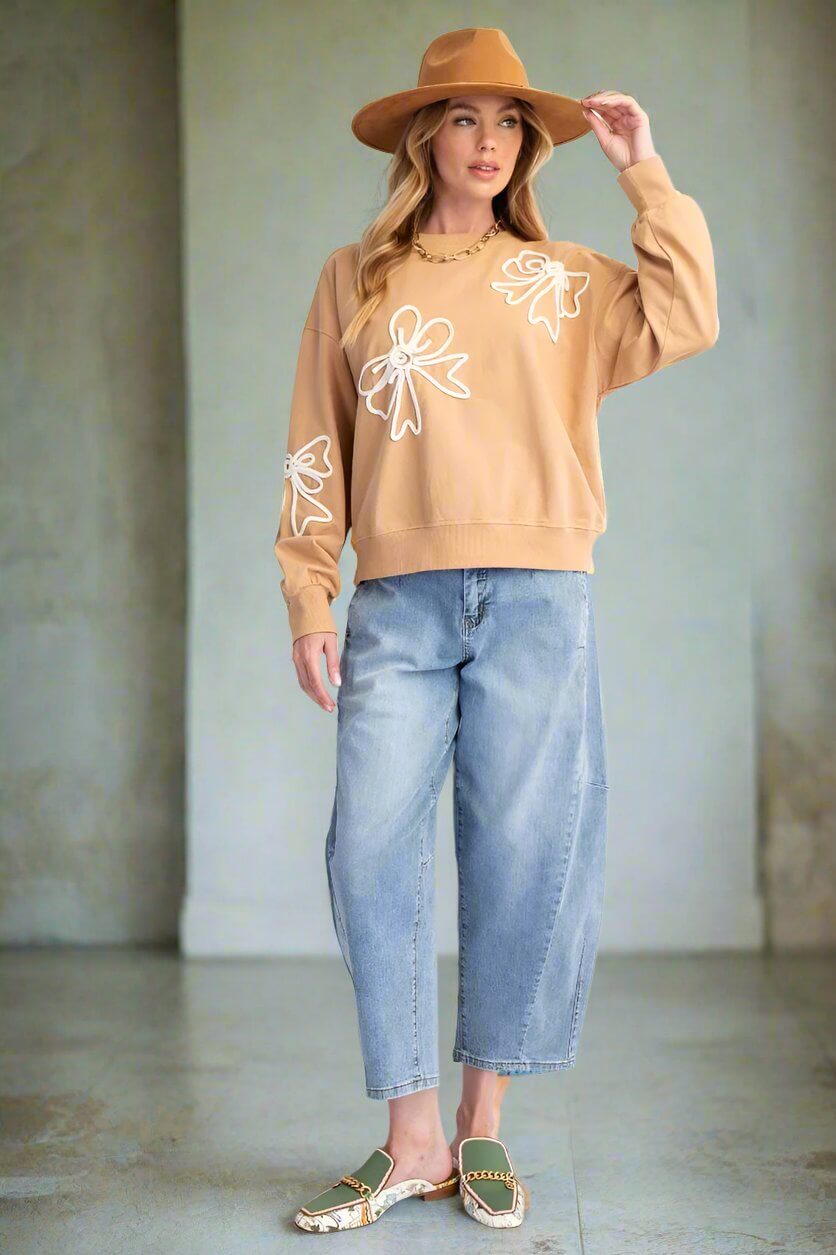 Woman wearing BOWS AND DREAMS Terracotta pullover with bow accents, relaxed fit, and wide-brim hat, paired with jeans and sandals.