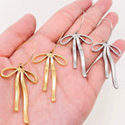 REFIEND RIBBON EARRINGS (2 colors) displayed in a hand, featuring gold and silver bow-inspired designs.