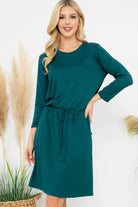 Woman wearing Emerald Enchantment Dress with 3/4 sleeves, cinched waist, and pockets. Comfortable knee-length, lightweight fabric.