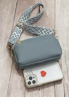 Light blue SKYE Crossbody Bag with a patterned strap next to a smartphone on a wooden surface.