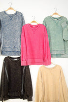 Five mineral-washed crew neck sweatshirts in gray, pink, green, black, and beige hanging on wooden hangers.