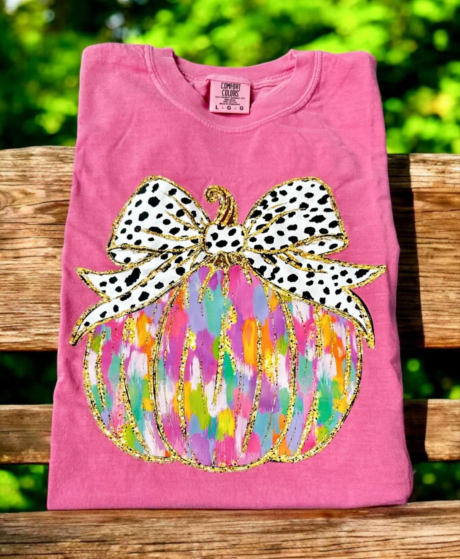 Crunchberry Pumpkin Art Tee by Comfort Color on a wooden bench, featuring a colorful pumpkin applique with a polka dot bow.