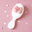 BOW BLISS MINI HAIRBRUSH on a pink background decorated with hearts, showcasing a cute 3D bow accent.