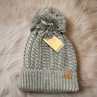 Taupe KNITTY GRITTY BEANIE by CC with cable knit, suede patch, and pom pom on a cozy beige background.