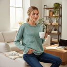 Casual woman in SKYLINE CREW (Dusty Teal) pullover, holding a mug in a bright, modern living room.