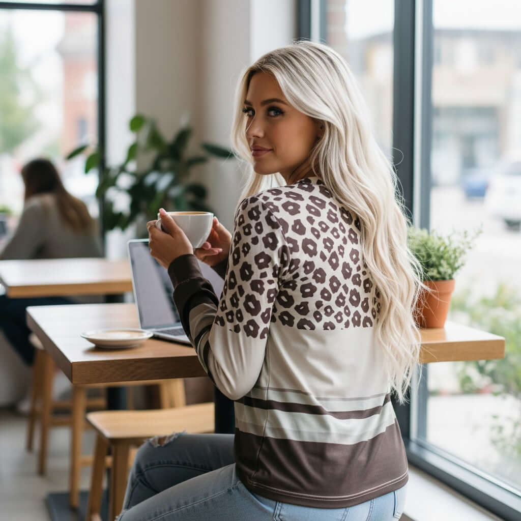 Woman wearing URBAN SAFARI TOP in a café, showcasing leopard color block design with coffee in hand.