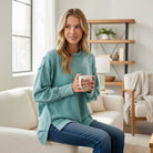 SOFT INTENTIONS Pullover by Zenana (dusty teal) worn by a woman holding a coffee mug, sitting in a cozy living room.