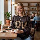 Woman wearing LOVE UNTAMED TOP - Easel with leopard patch letters, enjoying coffee in a cozy café setting.
