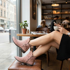 Woman wearing VERY G CAROLINA ANKLE BOOT in rose tan suede sitting in a café with stylish décor.