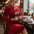 Woman wearing SPARKLE SEASON VELVET DRESS (Holiday Red) with rhinestone bow accents in a cozy cafe.