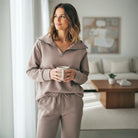 Model wearing QUIET LUXURY SET (taupe) with a cozy zip-collar top and jogger pants, enjoying a drink in a modern living space.