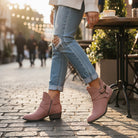 Woman wearing VERY G CAROLINA ANKLE BOOT in rose tan suede, styled with distressed jeans in an outdoor cafe.