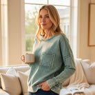 Woman wearing SKYLINE CREW (Dusty Teal) pullover, holding a cup in a bright room with cozy decor.