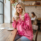 Woman wearing RIBBON ROMANCE SWEATER while enjoying coffee in a cozy café setting.