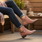 Woman wearing VERY G CAROLINA ANKLE BOOT in rose tan suede, styled with denim, showcasing side zip and buckle detail.