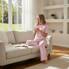 Woman relaxing in FESTIVE FRILLS LOUNGE SET, a pink comfy loungewear, sipping tea on a sofa in bright living room.