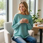 Model wearing the SOFT INTENTIONS Pullover by Zenana (dusty teal), holding a mug in a cozy café setting.