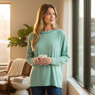 Woman wearing RELAX MODE TOP (sea green) holding a cup, showcasing its soft and relaxed style by a sunny window.