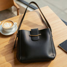Polished Path Handbag (black) on a wooden table next to a coffee cup, showcasing its sleek design and vegan leather construction.