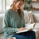 Model wearing SKYLINE CREW (Dusty Teal) reading a book in a cozy living room setting.