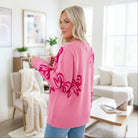 Woman wearing a RIBBON ROMANCE SWEATER with a pink bow pattern, enjoying coffee in a cozy living room.