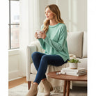 Woman in sea green RELAX MODE TOP enjoying a warm drink, sitting on a couch with a cozy atmosphere.
