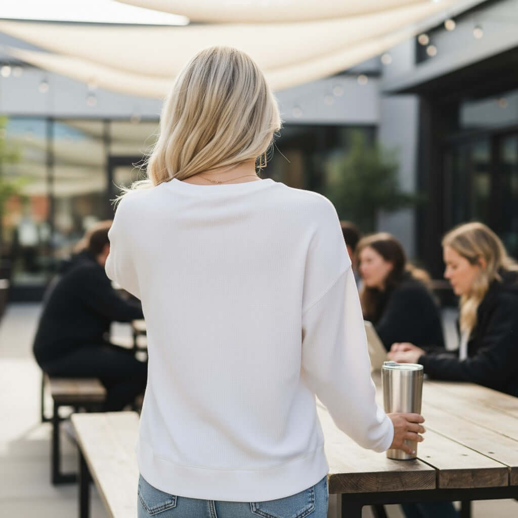 Model wearing the SOFTLY TIED CREWNECK white top with a bow print, showcasing a relaxed fit in a cafe atmosphere.