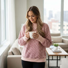 Model wearing the LAZY DAY LEOPARD CREWNECK, a pink leopard print sweatshirt, holding a coffee cup in a cozy living room.