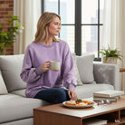 Woman wearing SOFT HORIZONS CREWNECK in lavender, sitting on sofa with coffee and breakfast.