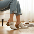 A person wearing HAZEL SLIP-ON MULES with a stylish knotted strap design and a block heel, sitting comfortably at home.