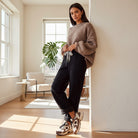 Woman wearing COMFORT CULTURE PANTS (BLACK) with a cozy sweater and stylish sneakers in bright indoor setting.
