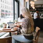 Woman wearing URBAN SAFARI TOP in a café, enjoying coffee and working on a tablet by the window.