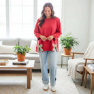 Model wearing SWEETHEART BOW TOP (red) with bow details, paired with relaxed jeans in a cozy living room setting.