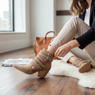Woman wearing ARISA STRAPPY ANKLE BOOTS in taupe with studded straps and block heel, sitting indoors.