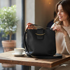 POLISHED PATH HANDBAG (black) displayed on a table next to coffee while a woman enjoys her drink.