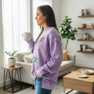 Woman in lavender MARDI GRAS STAR PULLOVER with sequined stars, holding a coffee mug in a stylish living room.