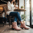 A woman wearing VERY G CAROLINA ANKLE BOOT in rose tan suede, sitting in a cozy café with a cup of coffee.