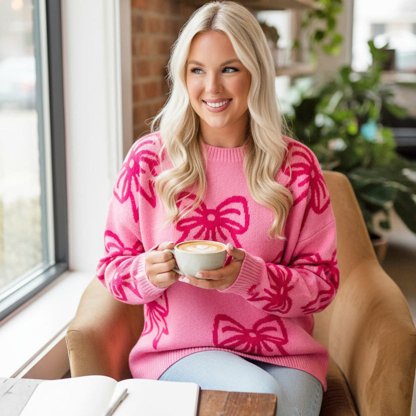 A woman wearing a Ribbon Romance Sweater with a pink bow pattern, enjoying coffee in a cozy setting.