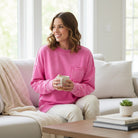 Woman enjoying a cozy moment wearing the CLOUD NINE CREW pullover in pink, seated on a sofa with a cup.