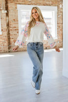 Woman showcasing the VANILLA SKYE TOP, a lightweight ribbed floral patchwork top, paired with flared jeans in a bright interior.