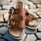 Feelin' Saucy sandals by Corkys with adjustable straps and shiny gold buckles on a cobblestone background.