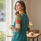 Woman wearing HIGHLAND RETREAT TOP with colorful plaid sleeves, enjoying coffee indoors.